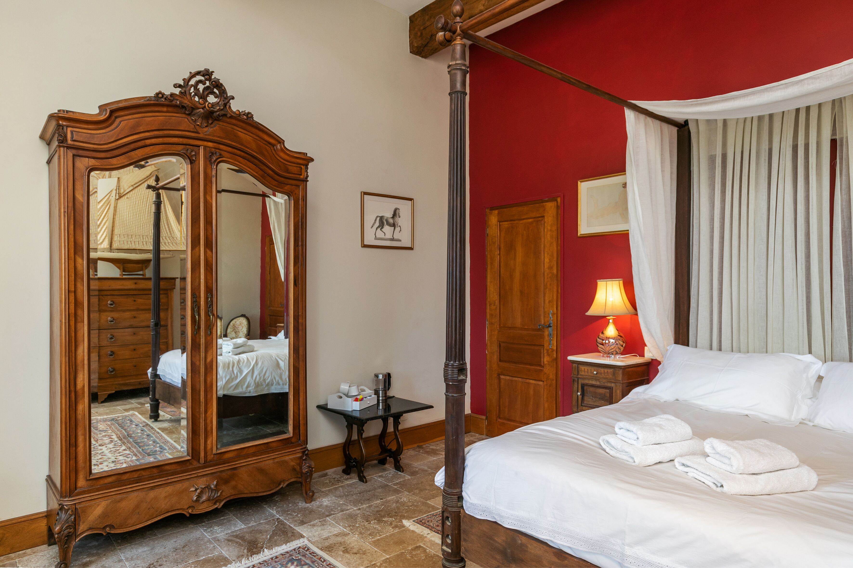 A bedroom with a four-poster bed, a wooden wardrobe, a bedside table with a lamp, and a mirror.