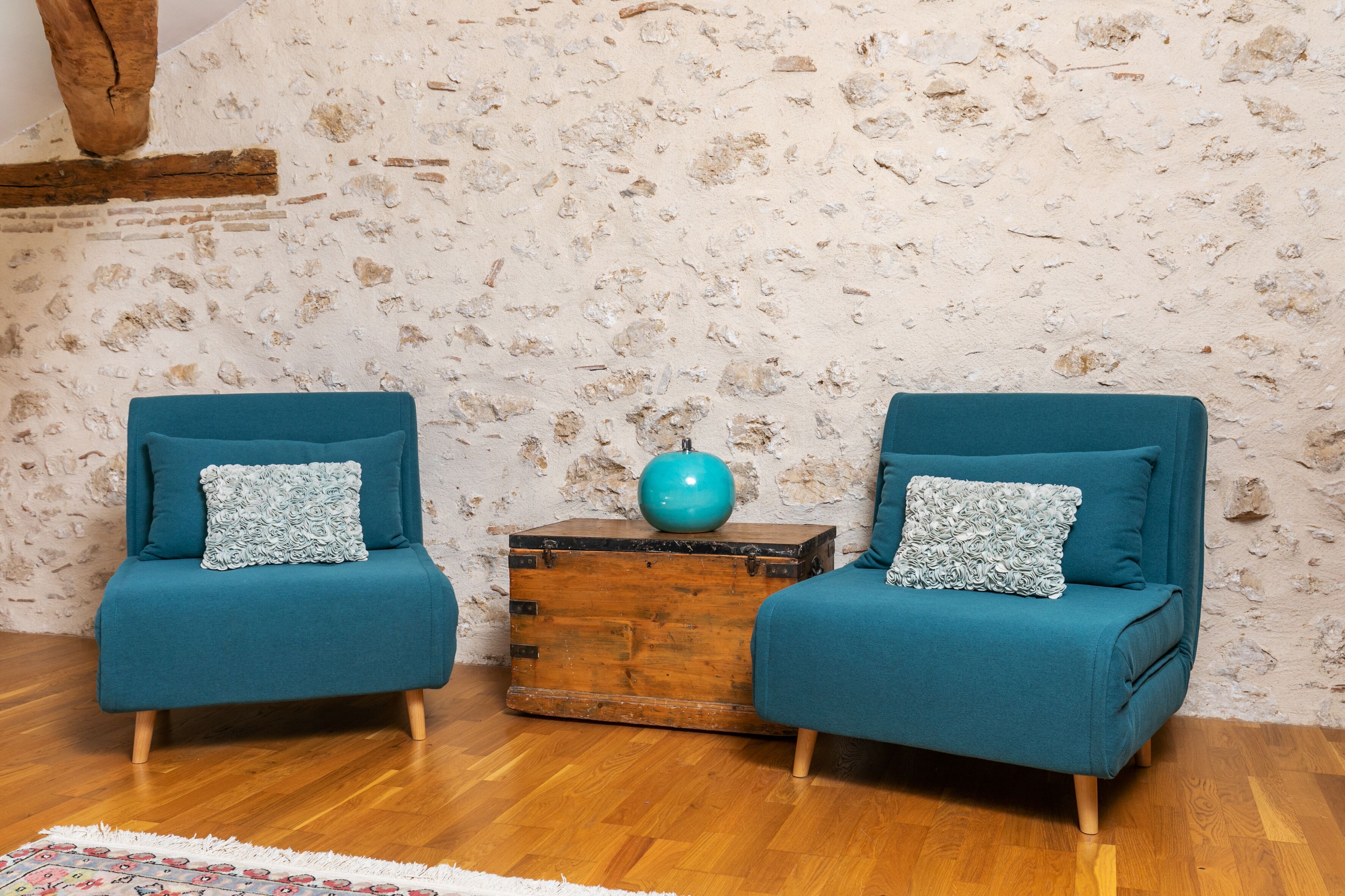 Two teal armchairs with patterned cushions, a wooden chest, and a globe on top, against a textured wall with exposed brick.