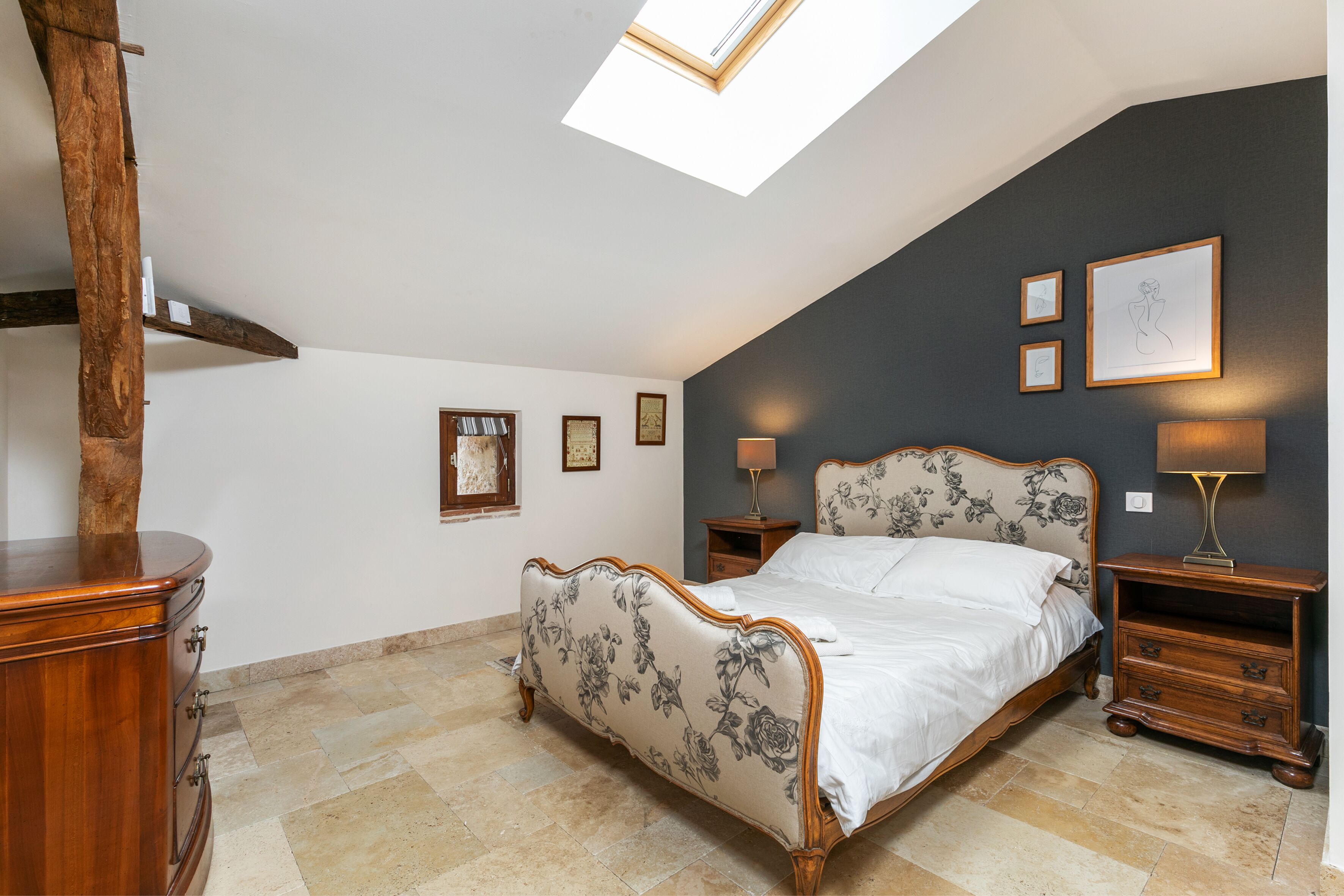 Un dormitorio con tragaluz, una cama con cabecera de flores, dos mesitas de noche con lámparas y un aparador de madera.
