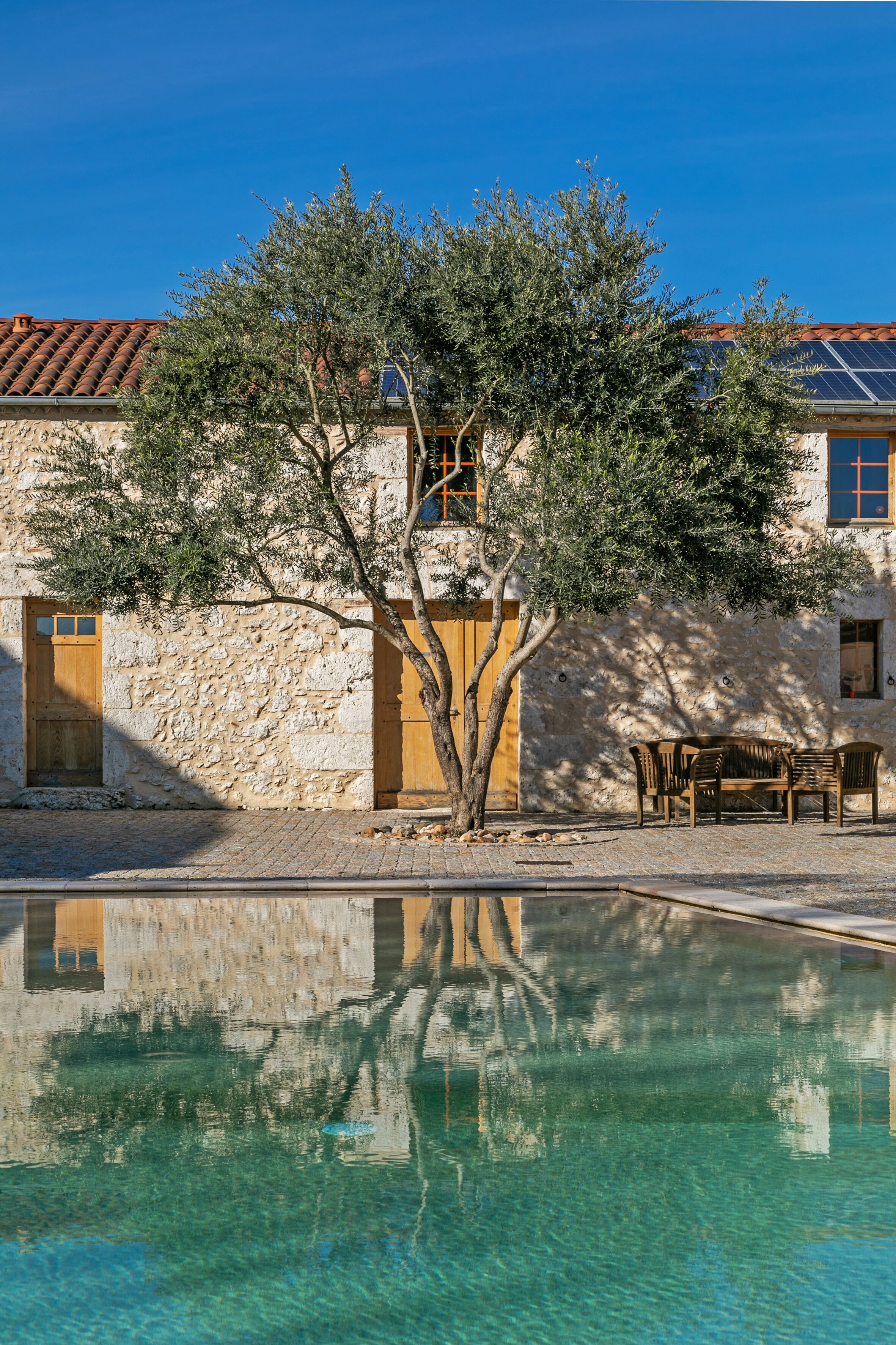 A stone building with a tiled roof, a swimming pool, and a tree in front.
