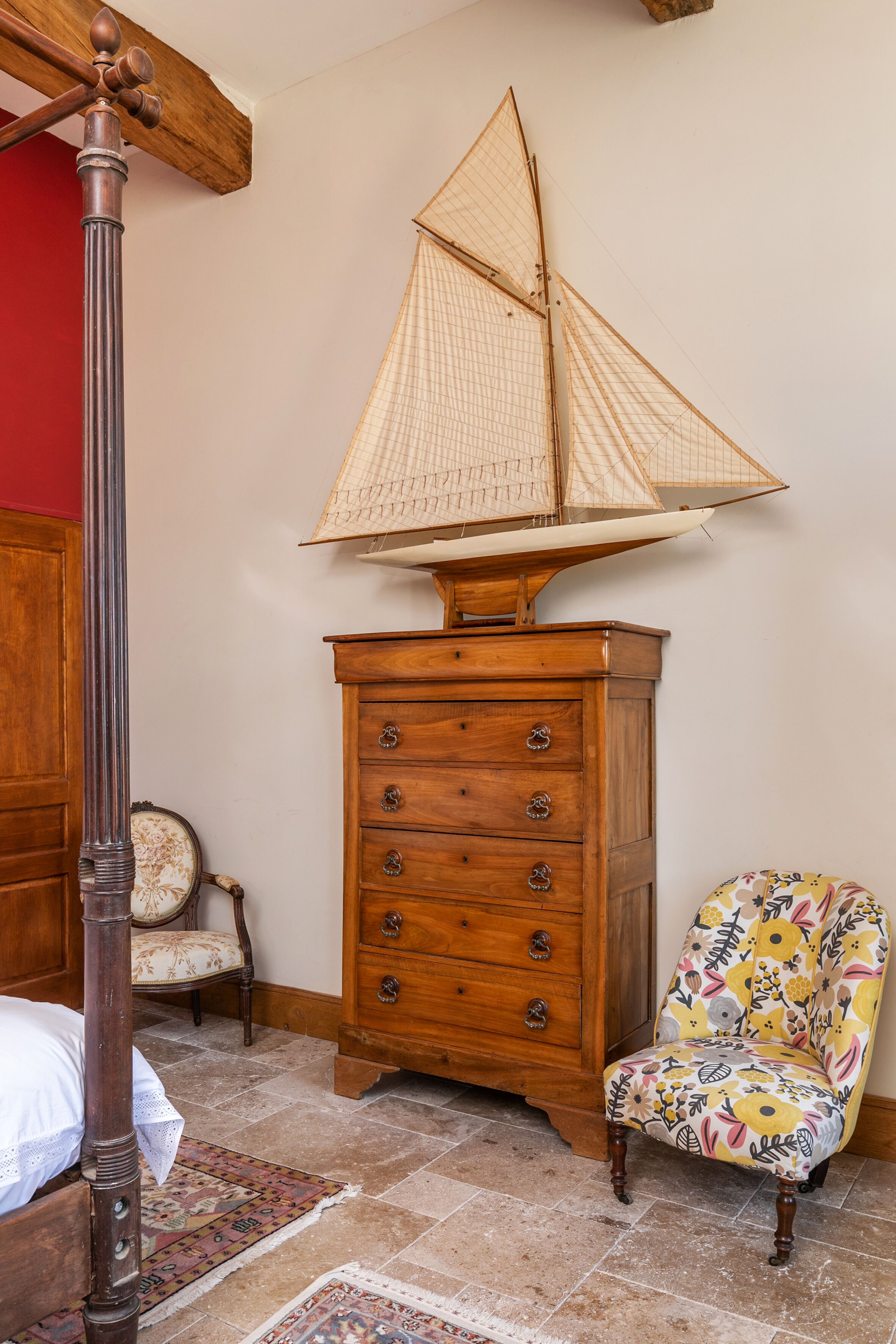 A bedroom with a wooden four-poster bed, a wooden chest of drawers, a floral armchair, and a ship-shaped lamp.