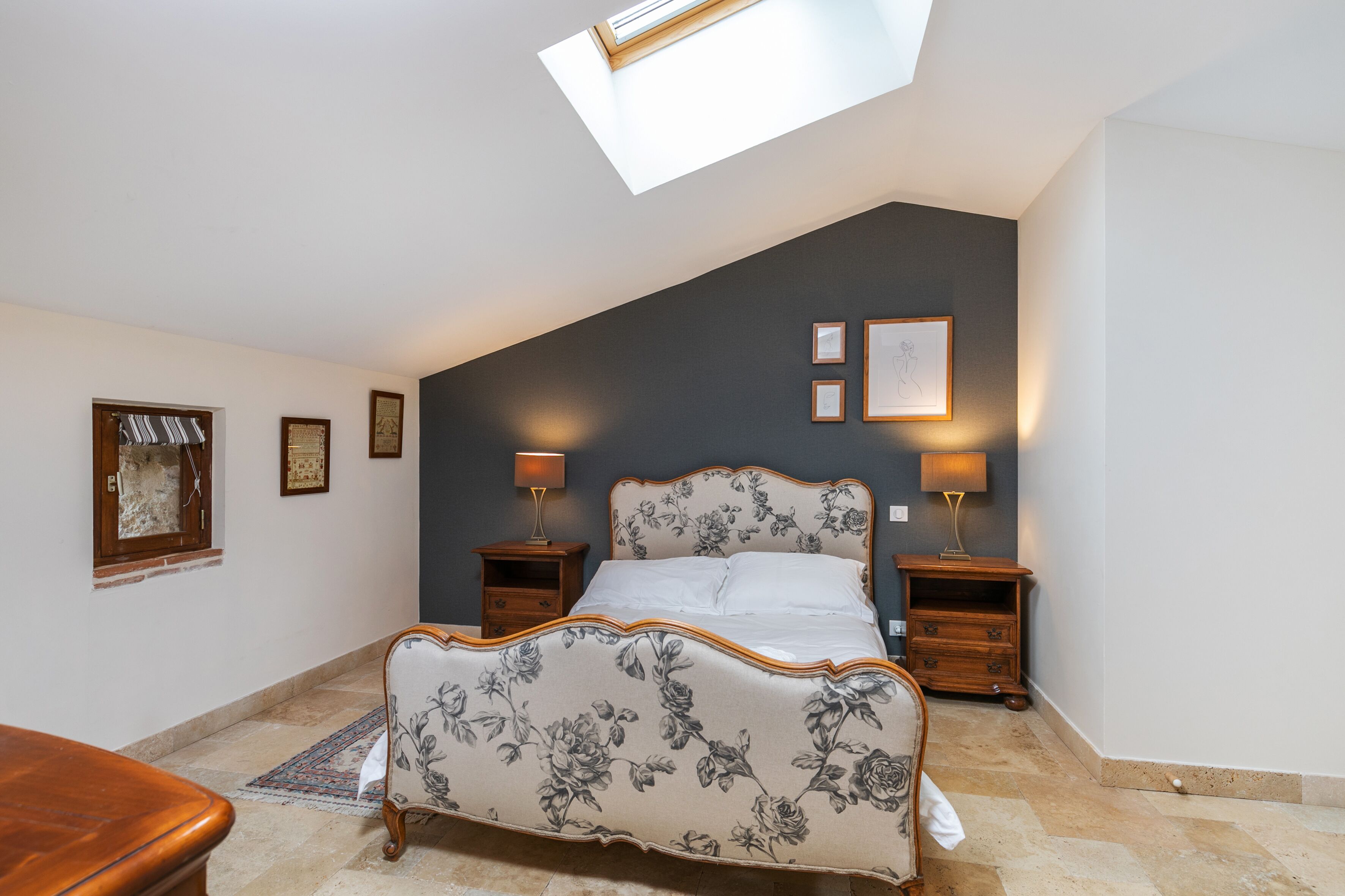 A bedroom with a sloped ceiling, a bed with floral upholstery, two bedside tables with lamps, and framed pictures on the wall.