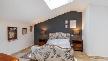 A bedroom with a sloped ceiling, a bed with floral upholstery, two bedside tables with lamps, and framed pictures on the wall.