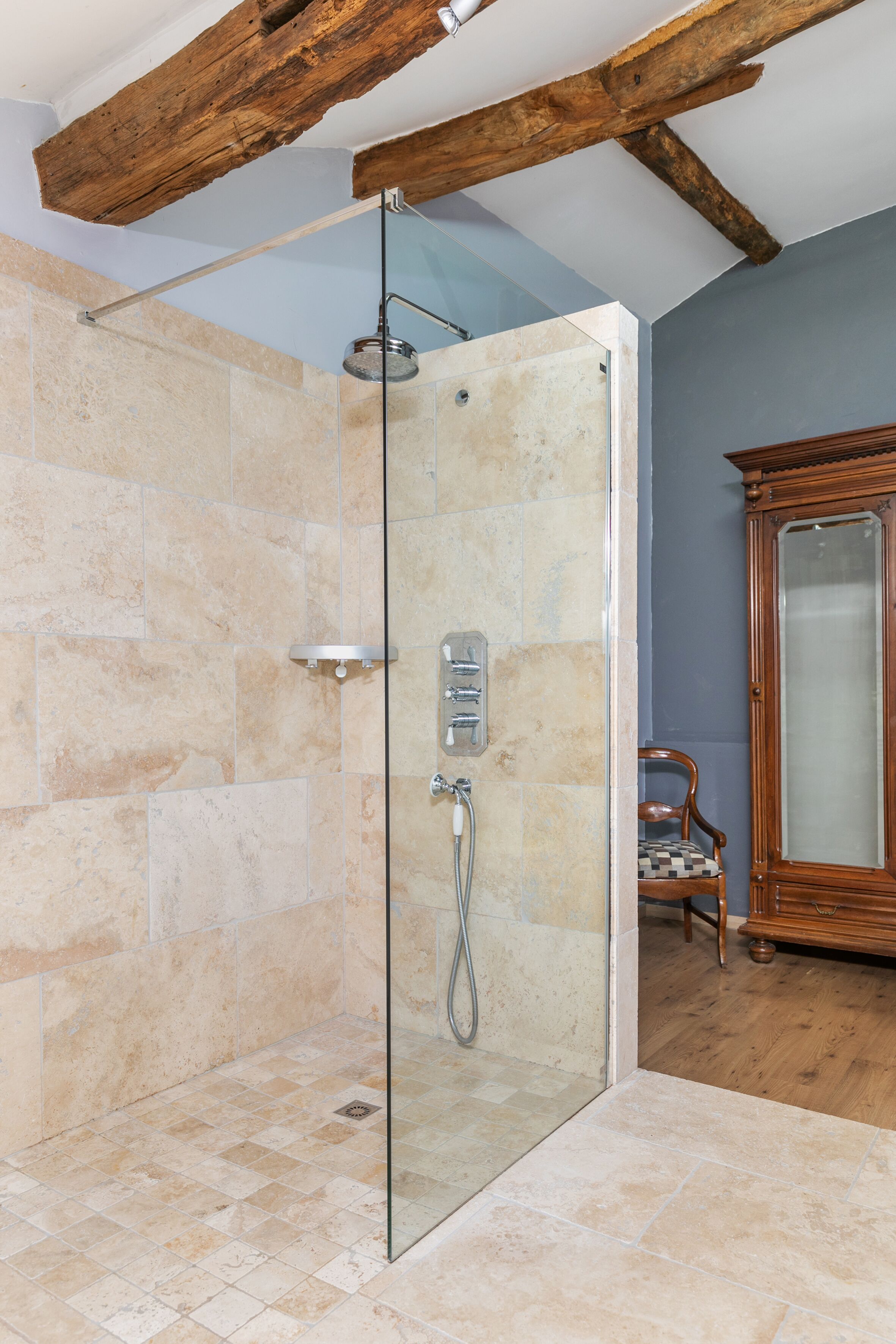 A glass-enclosed shower with stone tiles, a wooden chair, and a cabinet.
