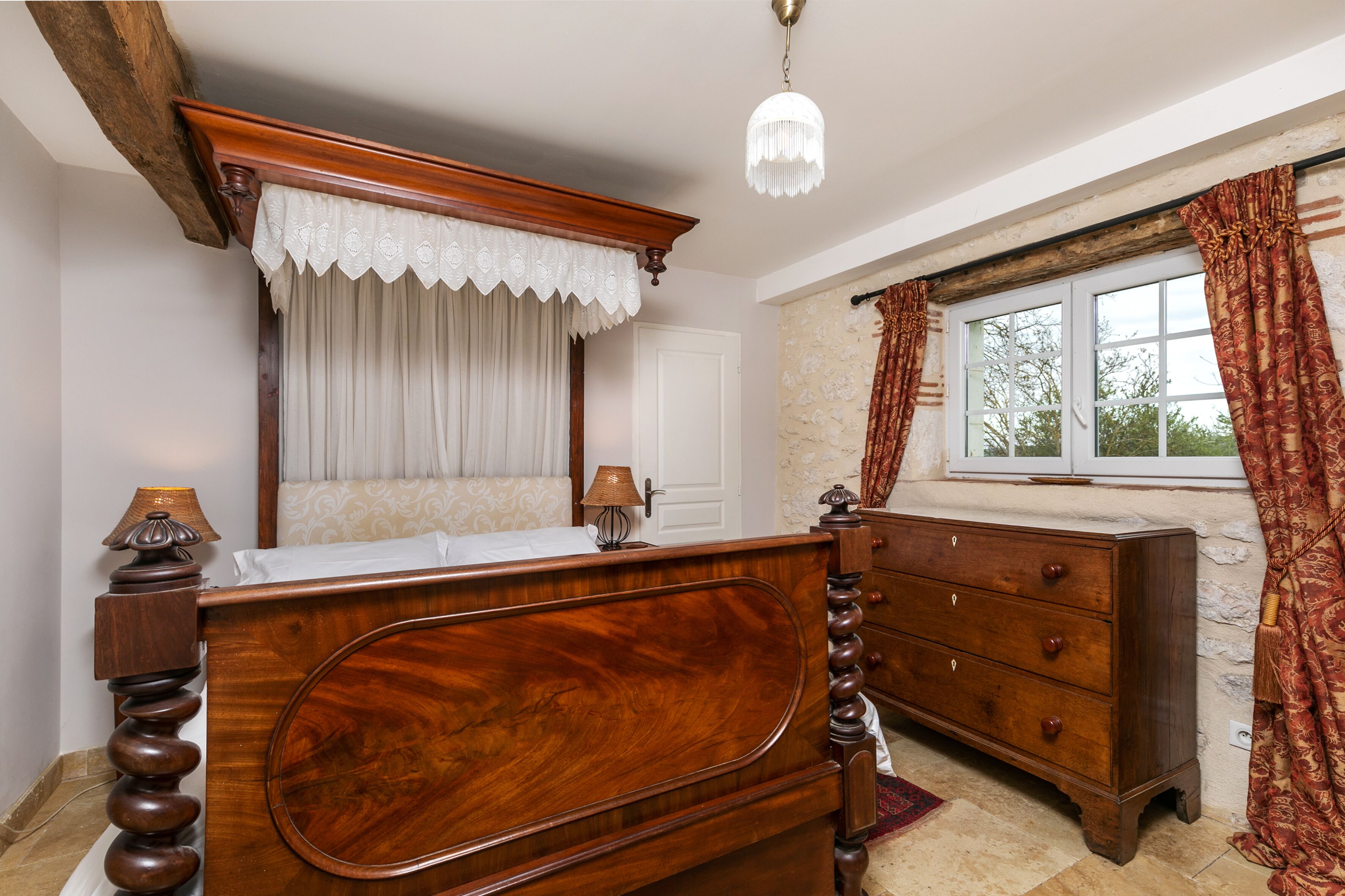 Un dormitorio con cama de madera, cómoda, ventana con cortinas y un candelabro.