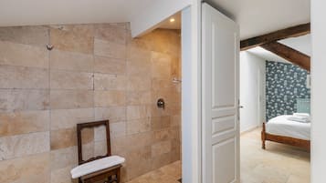 Une salle de bain carrelée de carreaux beiges, avec un banc en bois et une porte donnant sur une chambre à coucher équipée d’un lit et ornée d’un papier peint à motifs floraux.