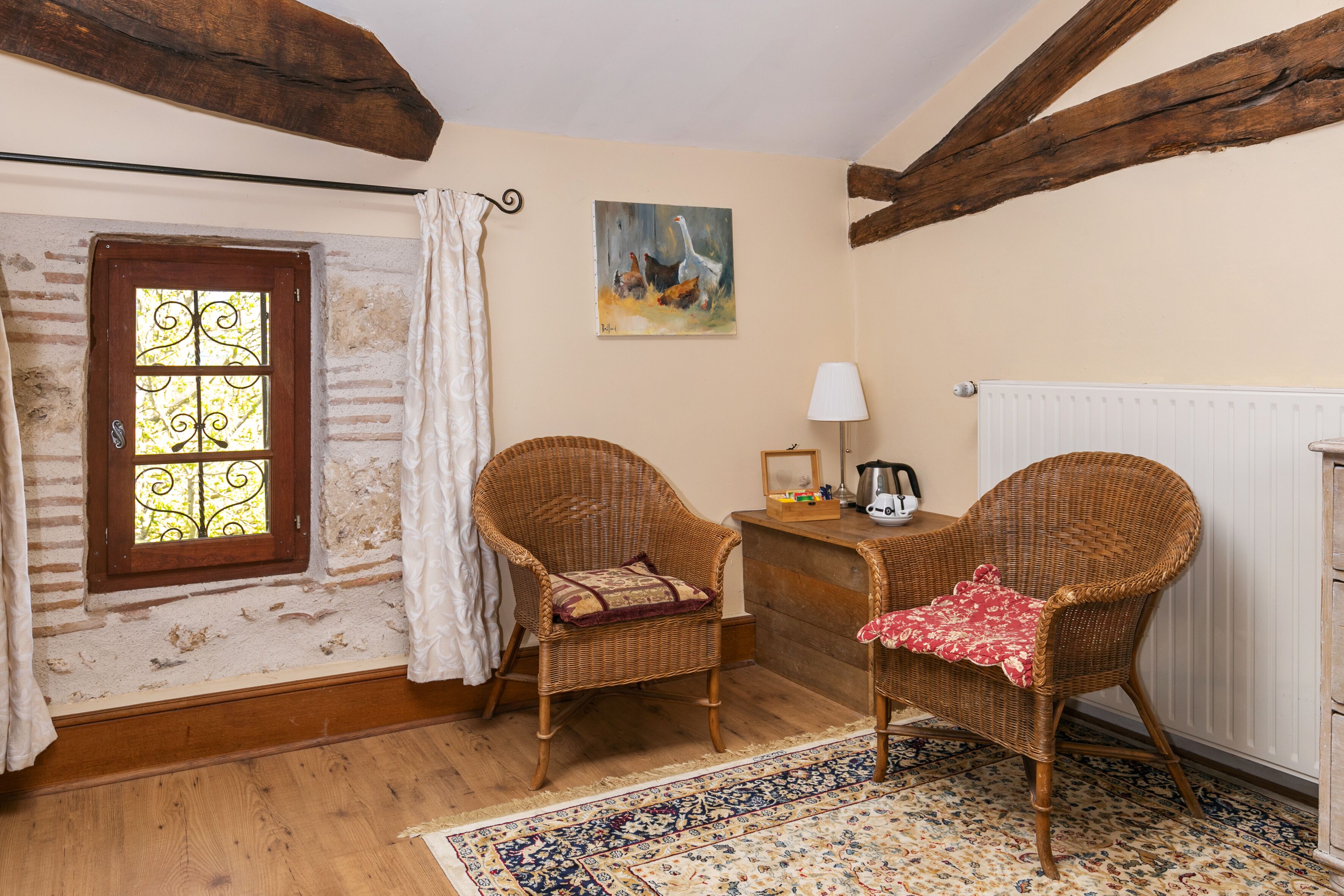 A cozy room with two wicker chairs, a wooden floor, a patterned rug, a radiator, and a window with curtains.