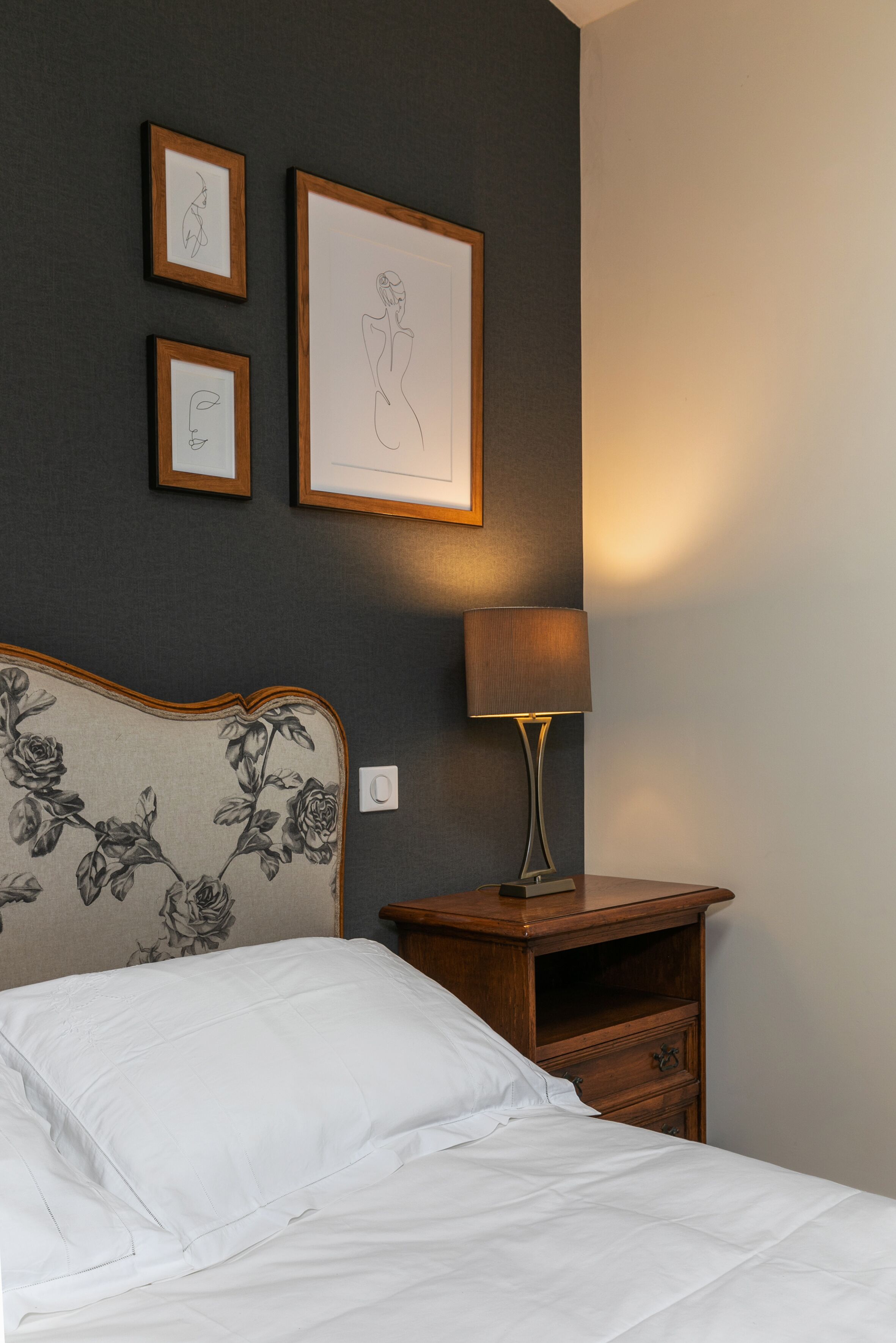 A neatly made bed with white linens, a wooden nightstand with a lamp, and three framed artworks on the wall.