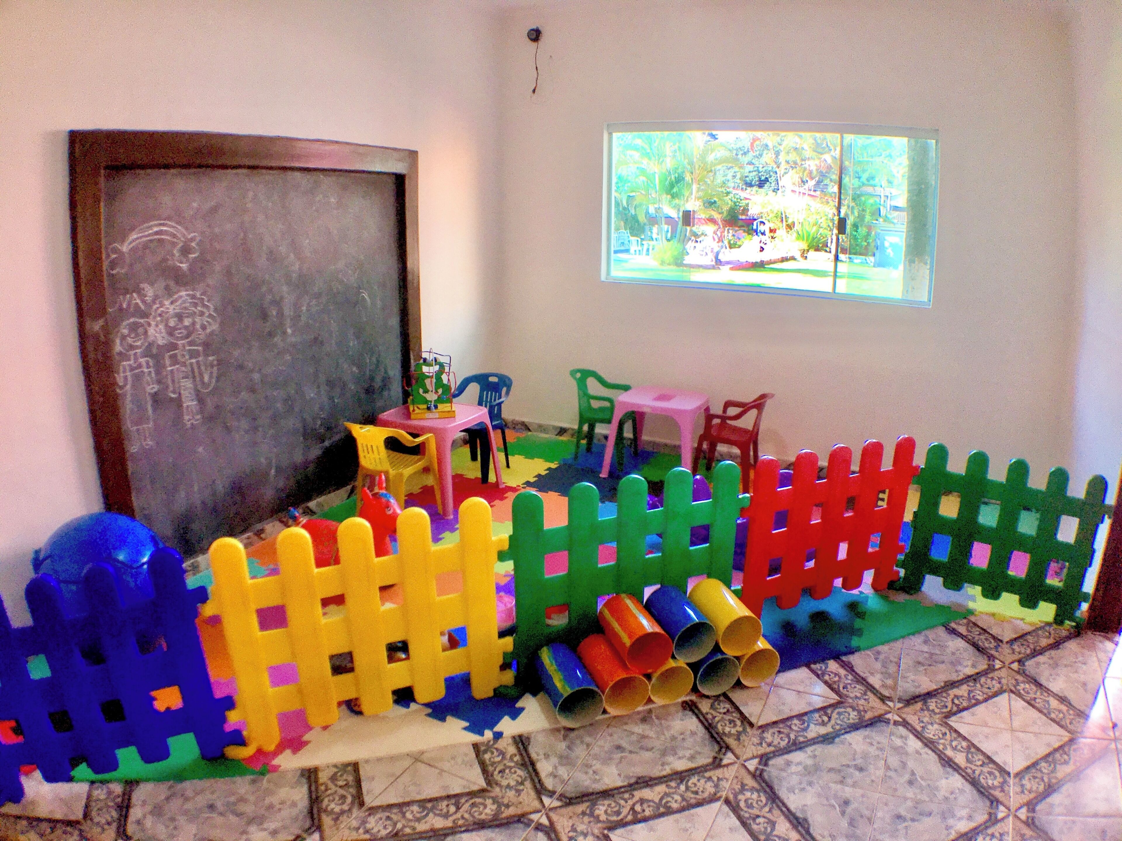 Una sala de juegos con un corralito colorido, una mesita pequeña, sillas y una pizarra con dibujos.