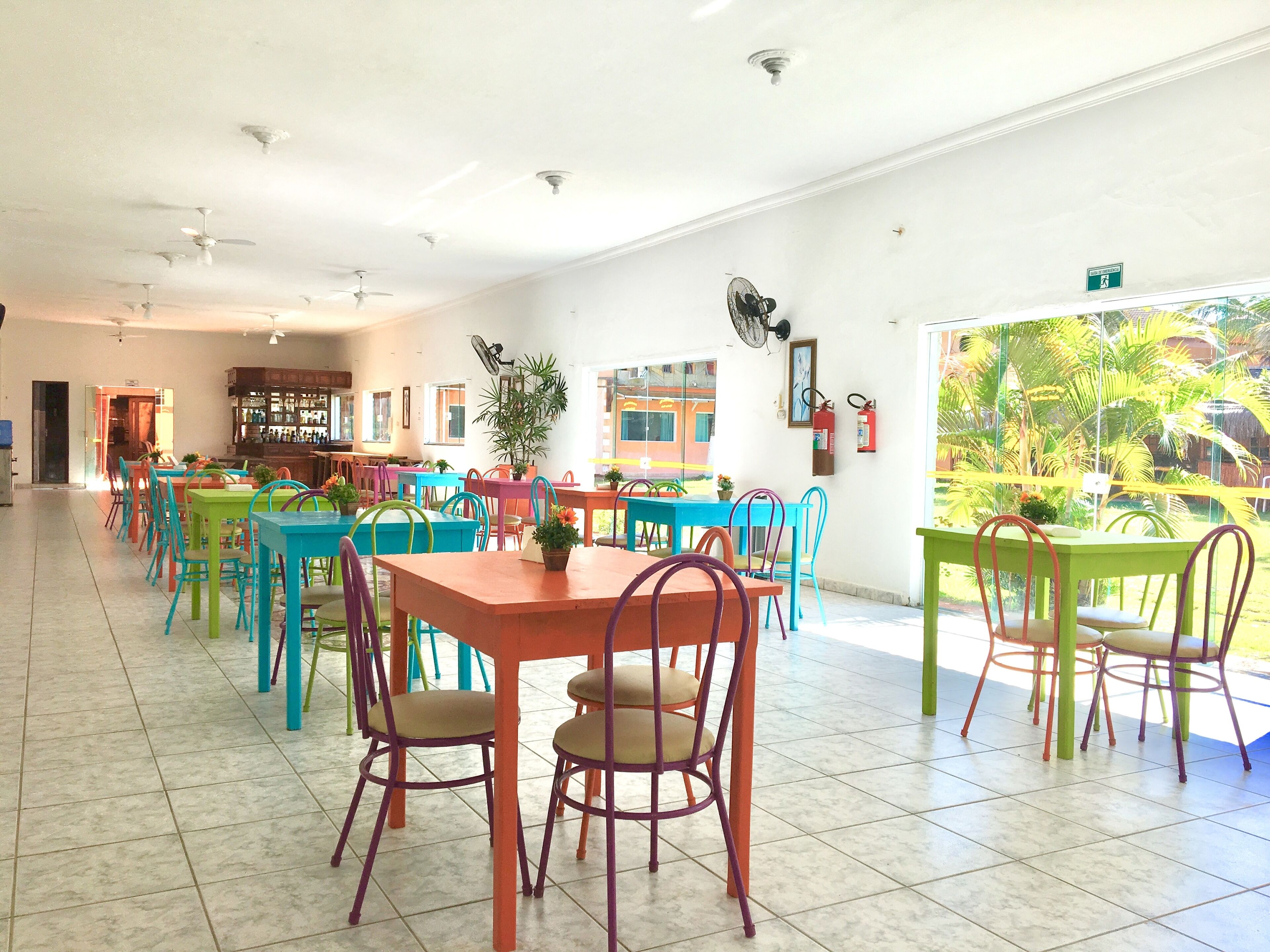 A spacious indoor area with multiple colorful tables and chairs, a bar area, and a view of greenery outside.
