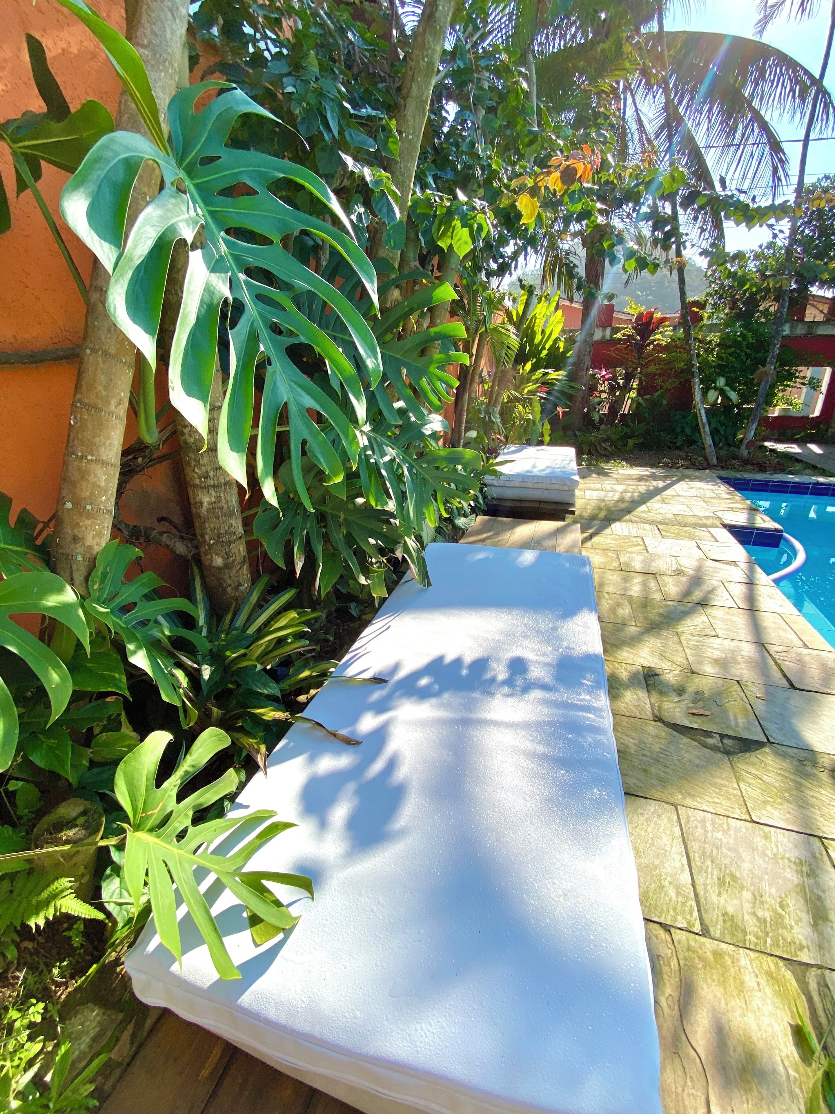 A poolside area with a white lounge chair, surrounded by lush tropical plants and a clear blue pool.