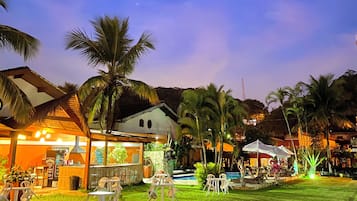 Área externa bem iluminada, com piscina, palmeiras e um edifício com telhado de palha.
