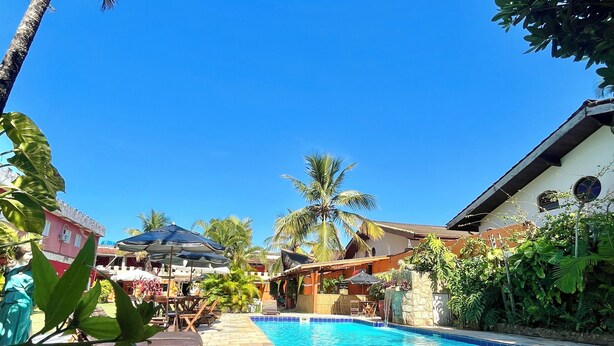 A swimming pool with clear blue water, surrounded by lush greenery and a clear blue sky. - Pousada Dolce Vita (Itanhaém)