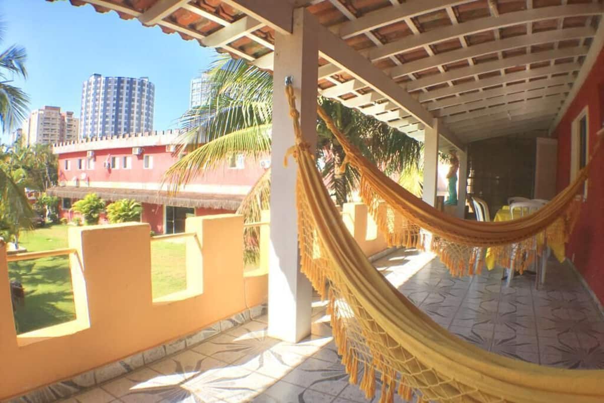 A covered outdoor area with a hammock, tiled floor, and a view of a building and palm trees.