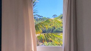 A room with a bed, white curtains, and a view of a tropical landscape through a window. - Pousada Dolce Vita (Itanhaém)