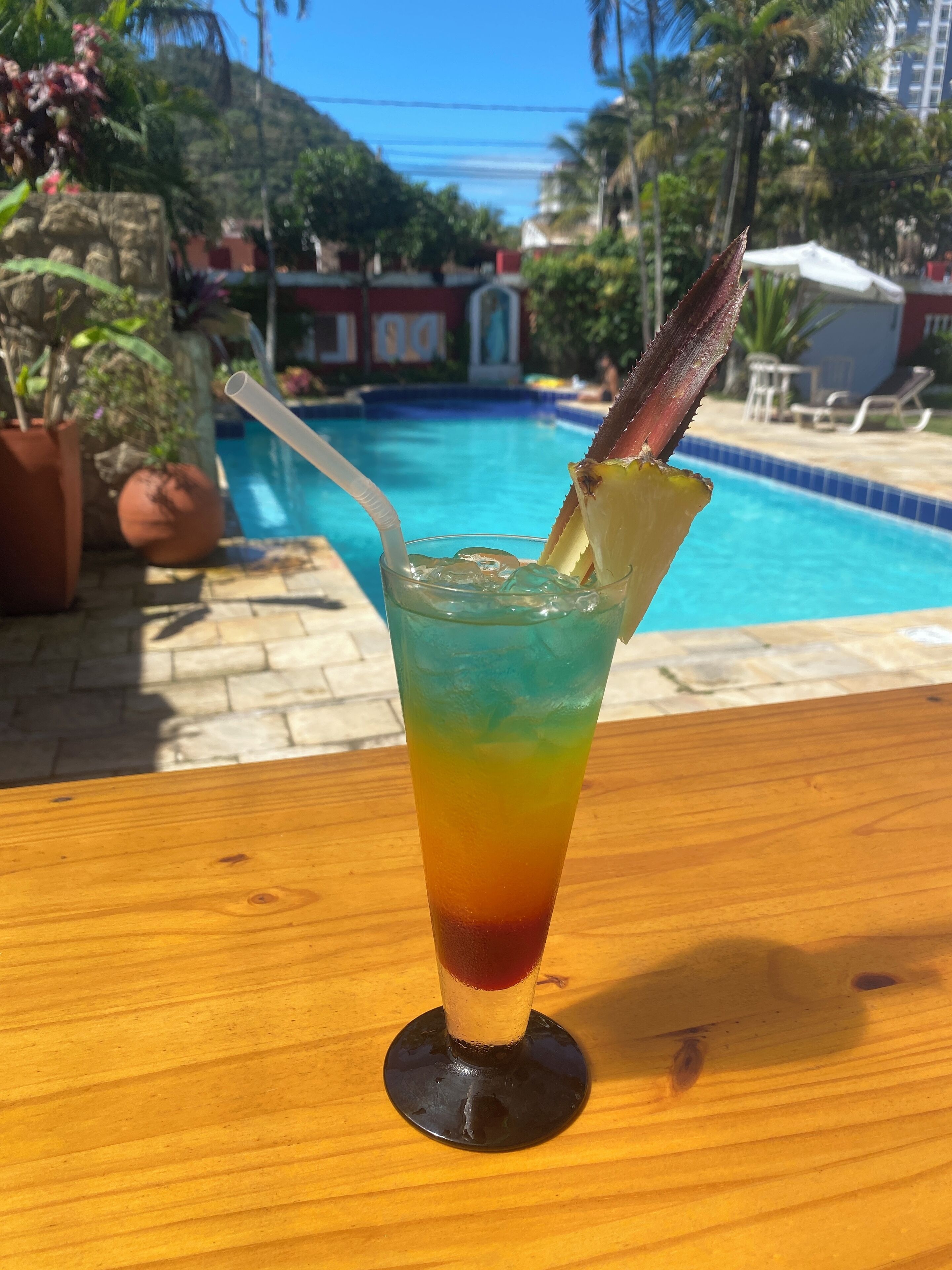 A glass of colorful drink with a straw and a piece of fruit on a wooden table by a pool.