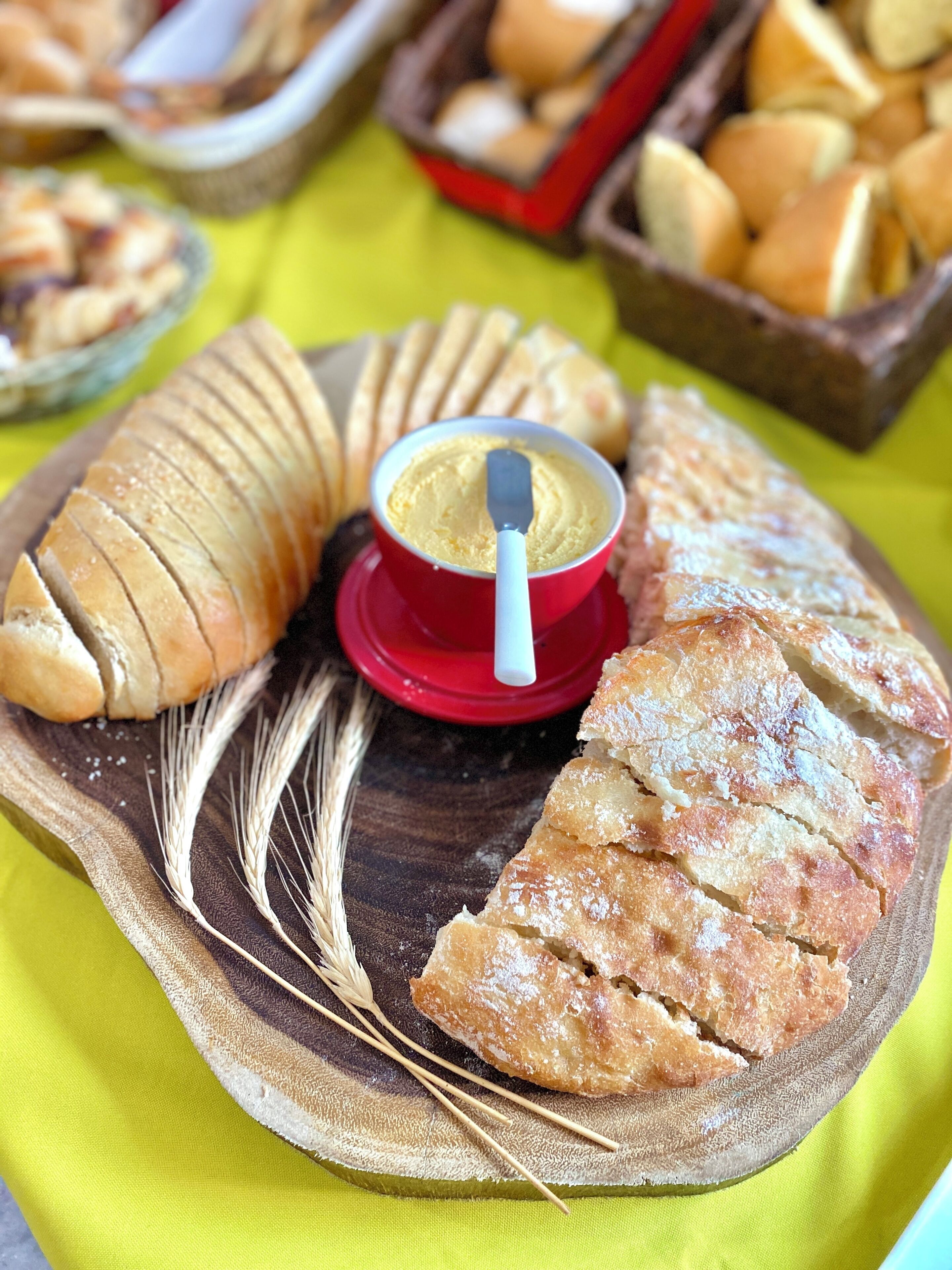 A wooden serving board with sliced bread, a bowl of butter, and wheat stalks.