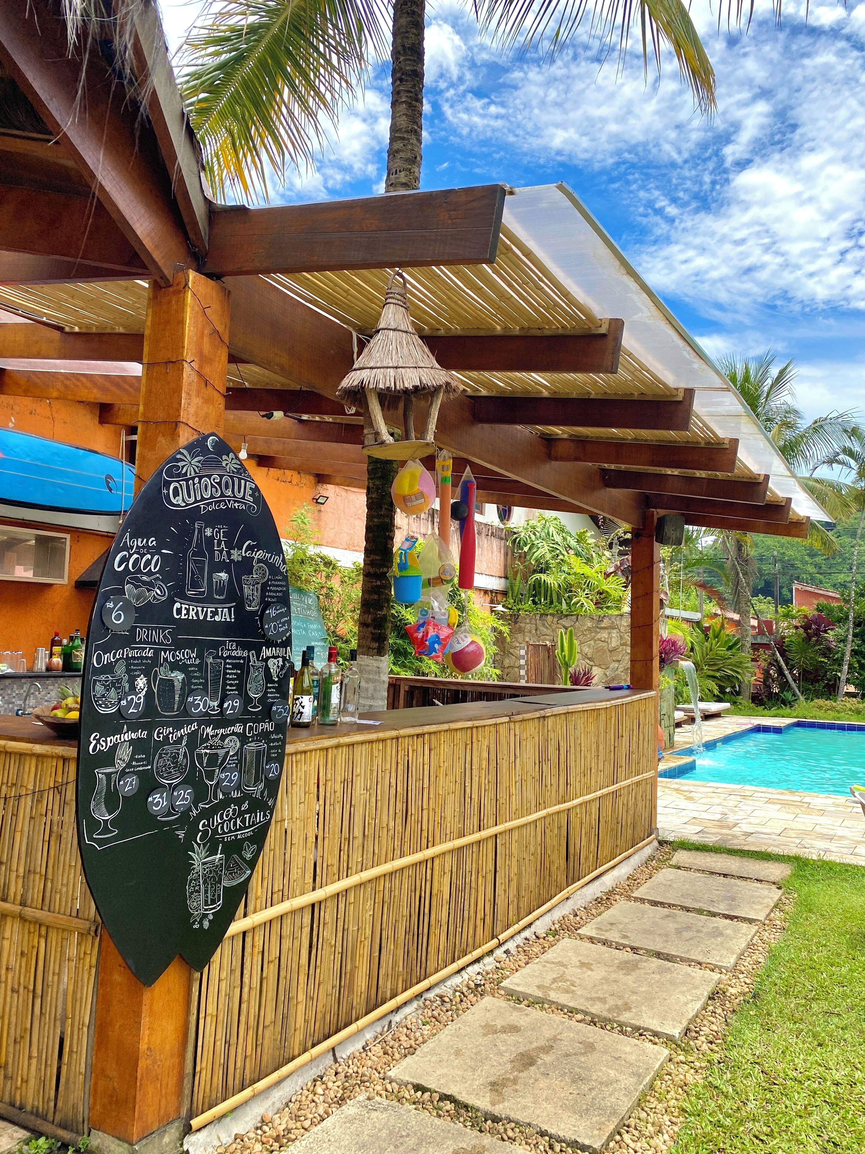Zona de bar con un menú en la pizarra que enumera bebidas y cócteles, una piscina y un entorno tropical al aire libre.
