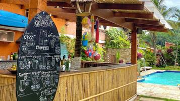 Zona de bar con un menú en la pizarra que enumera bebidas y cócteles, una piscina y un entorno tropical al aire libre.