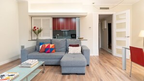 Una sala moderna con un sofá seccional gris, una mesita de centro pequeña y una estantería. Se ve una cocina al fondo con gabinetes rojos y una ventana con contraventanas blancas.