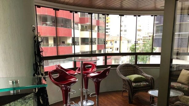 A balcony with a glass table and red chairs, a wicker chair, and a view of a red and white building. - Apto Perfeito Quadra Mar (Balneário Camboriú)