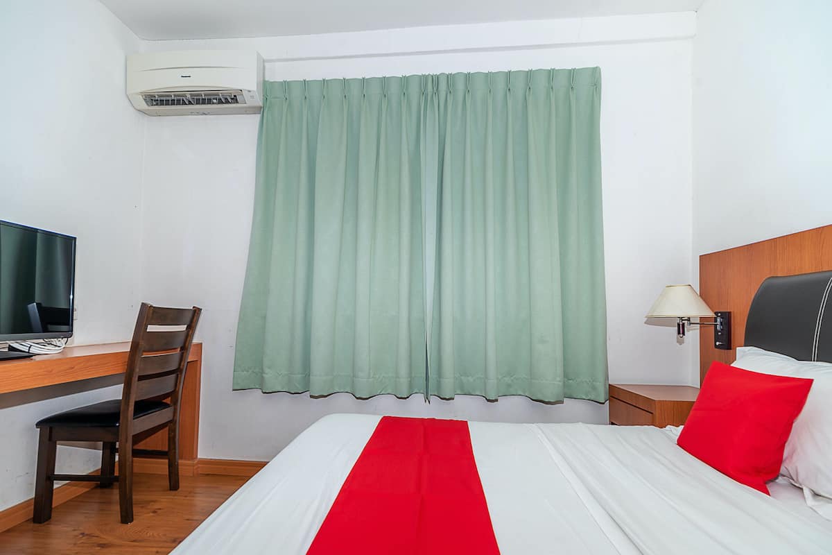 Habitación de hotel con una cama, un escritorio con computadora, una silla, una lámpara y una ventana con cortinas.