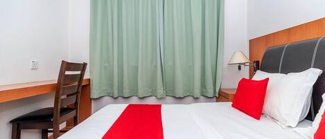 Une chambre d’hôtel avec un lit, un bureau, une chaise, une lampe et une fenêtre avec des rideaux.