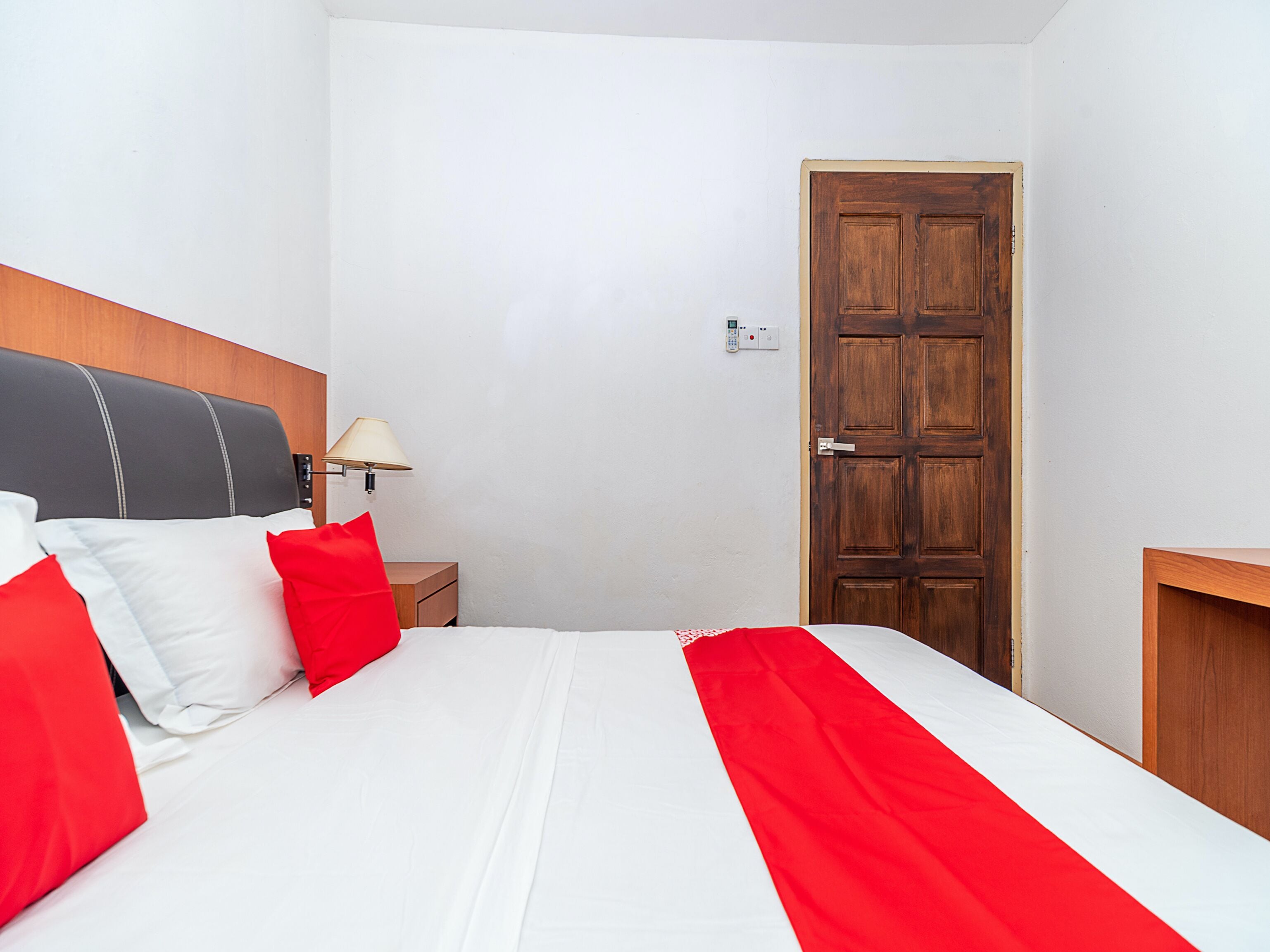 A hotel room with a bed, a wooden door, a nightstand, and a wall-mounted lamp.