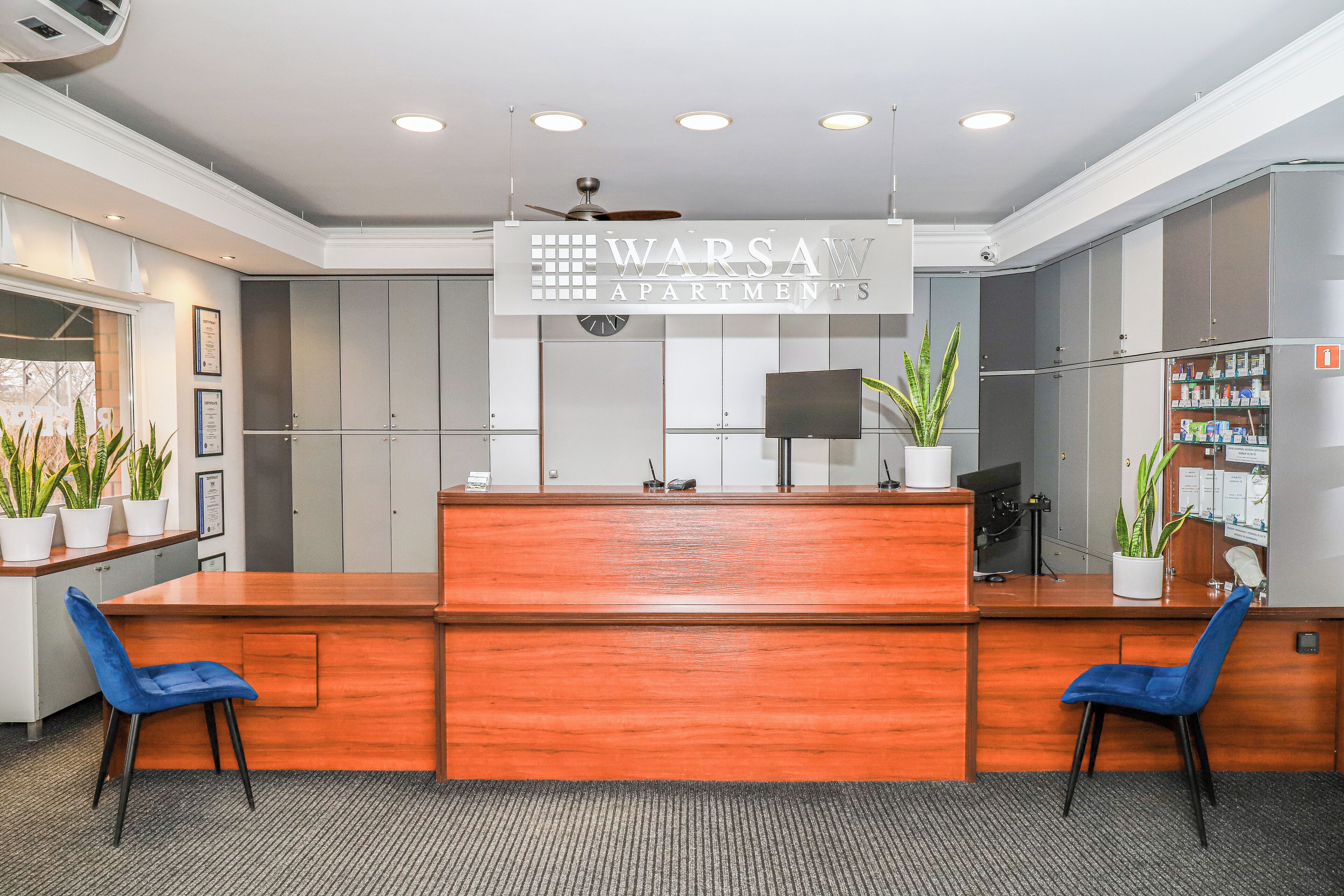 A reception area with a wooden desk, two blue chairs, a ceiling fan, and a sign for Warsaw Apartments.