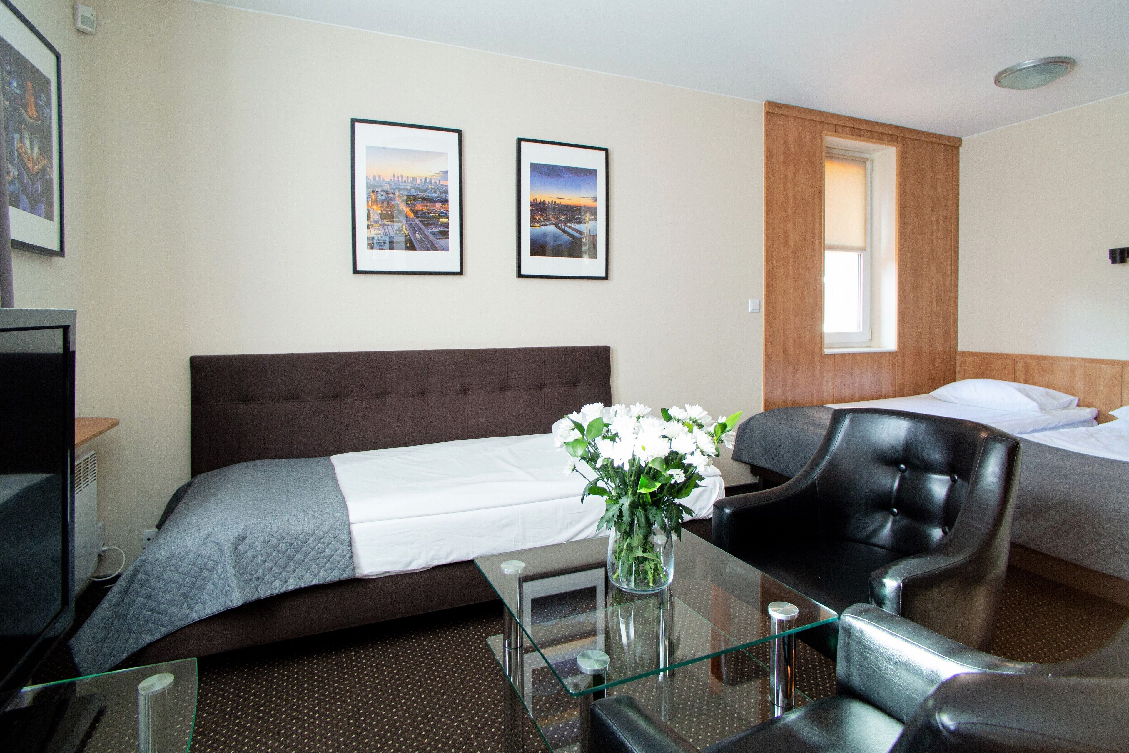 Habitación de hotel con una cama, una silla, un escritorio con un florero y dos cuadros en la pared.