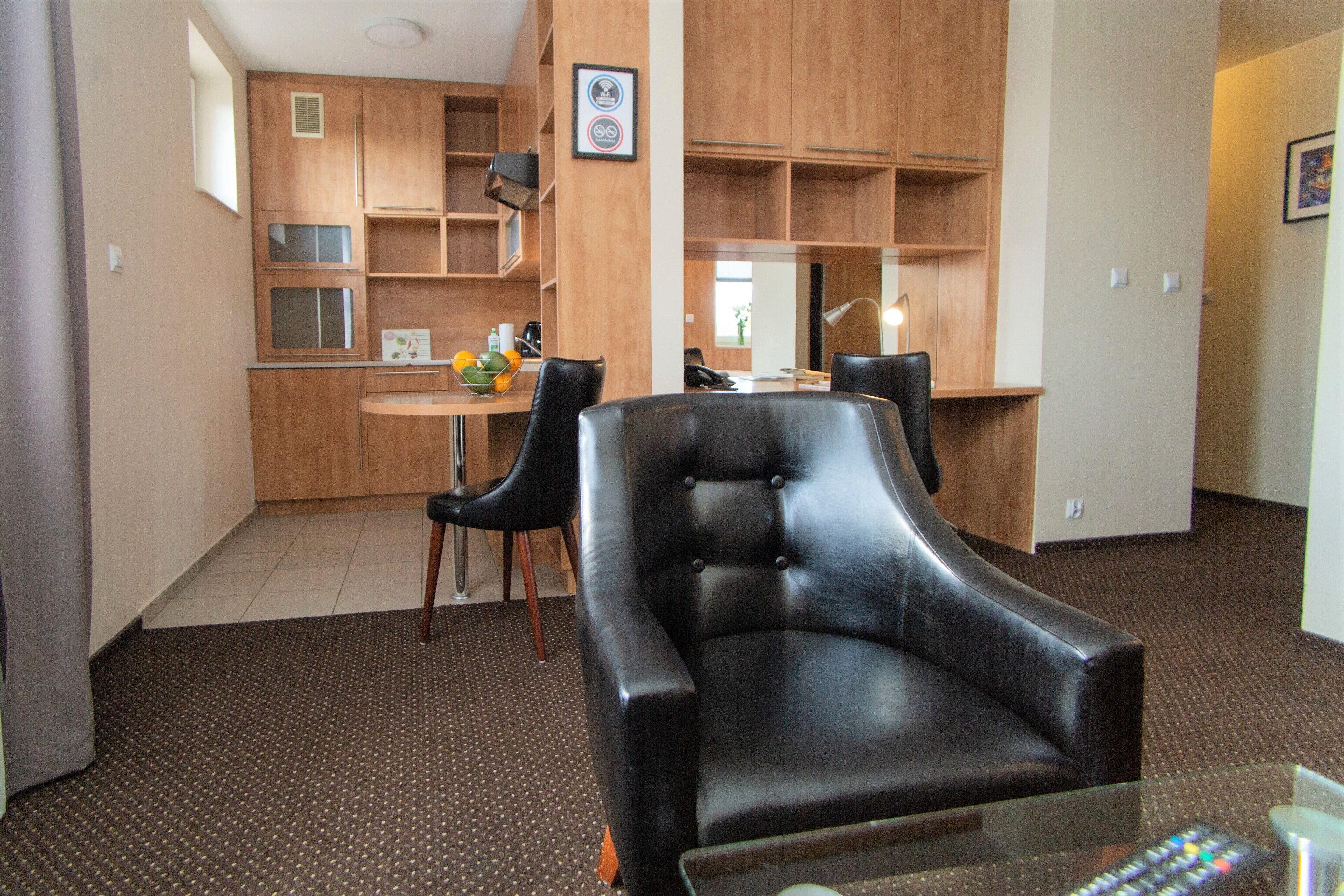 A living room with a black leather sofa, a glass coffee table, and a kitchen area with wooden cabinets and a dining table.