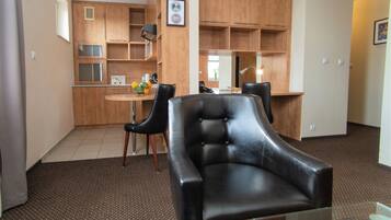 A living room with a black leather sofa, a glass coffee table, and a kitchen area with wooden cabinets and a dining table.