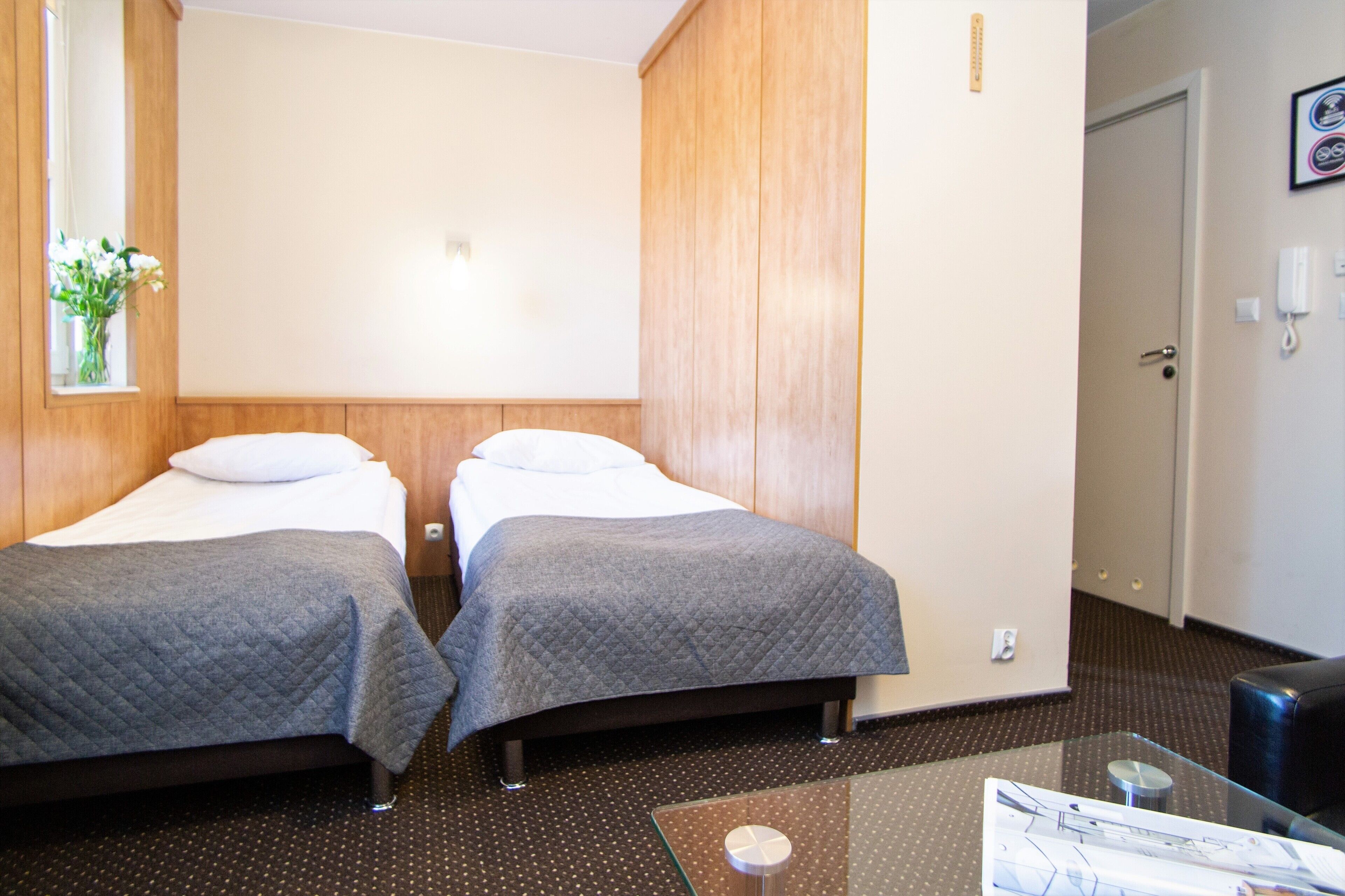 A hotel room with two beds, a glass table, and a small window with flowers.