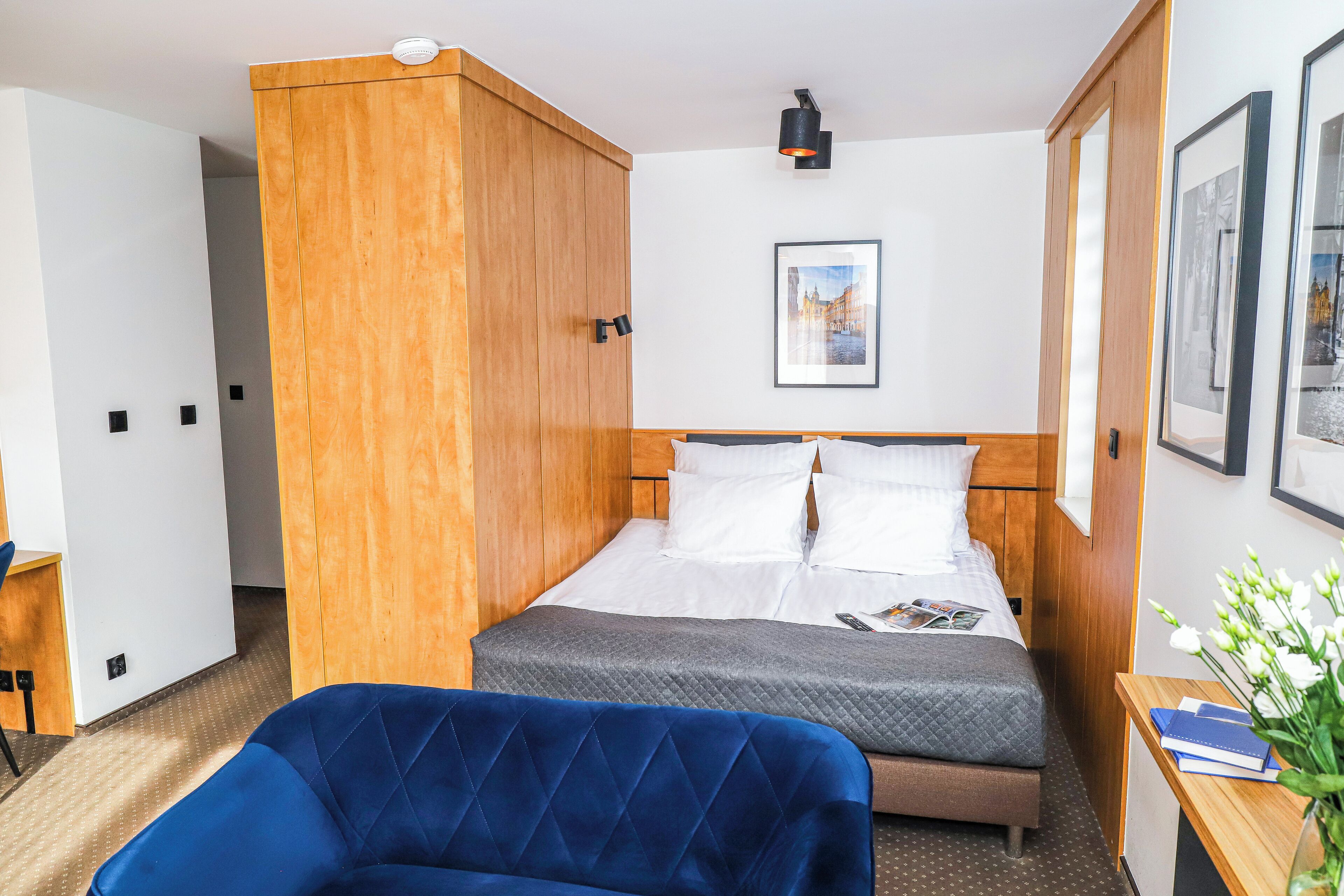 Habitación de hotel con una cama, una silla azul, un armario de madera, una mesita de noche con un libro y un jarrón de flores, y un cuadro en la pared.