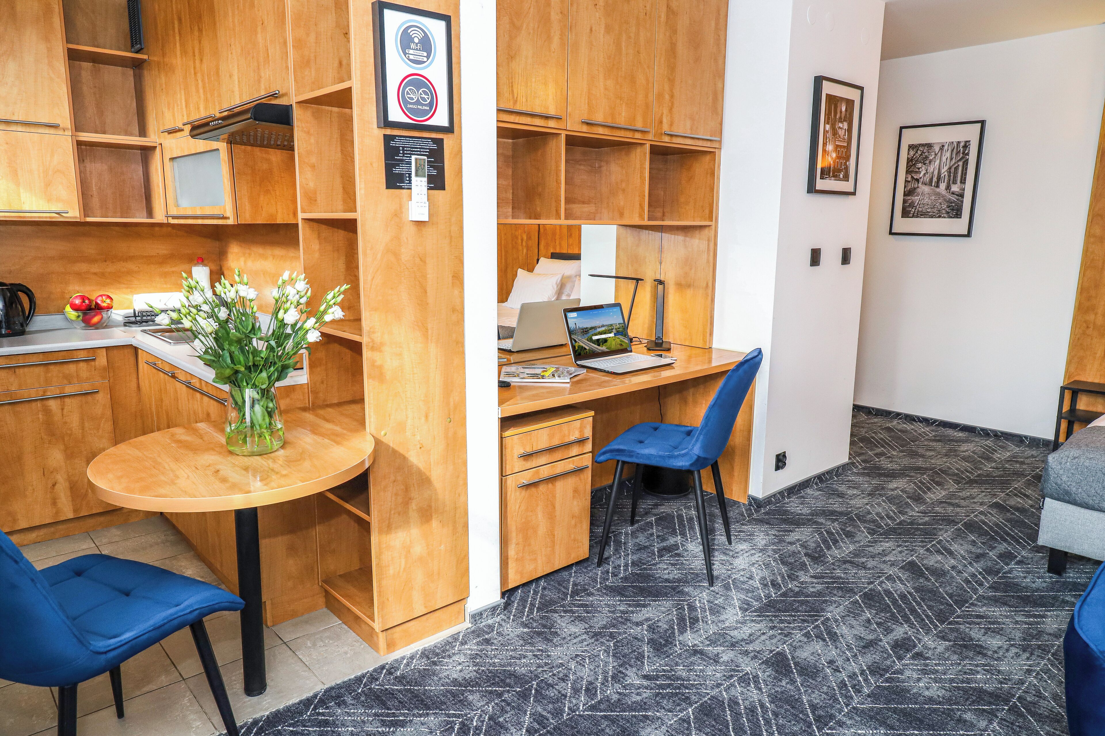 A compact room with a wooden desk, a laptop, a chair, a small table with a vase of flowers, and a kitchenette area.