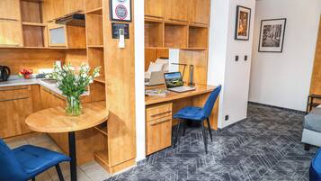 A compact room with a wooden desk, a laptop, a chair, a small table with a vase of flowers, and a kitchenette area.