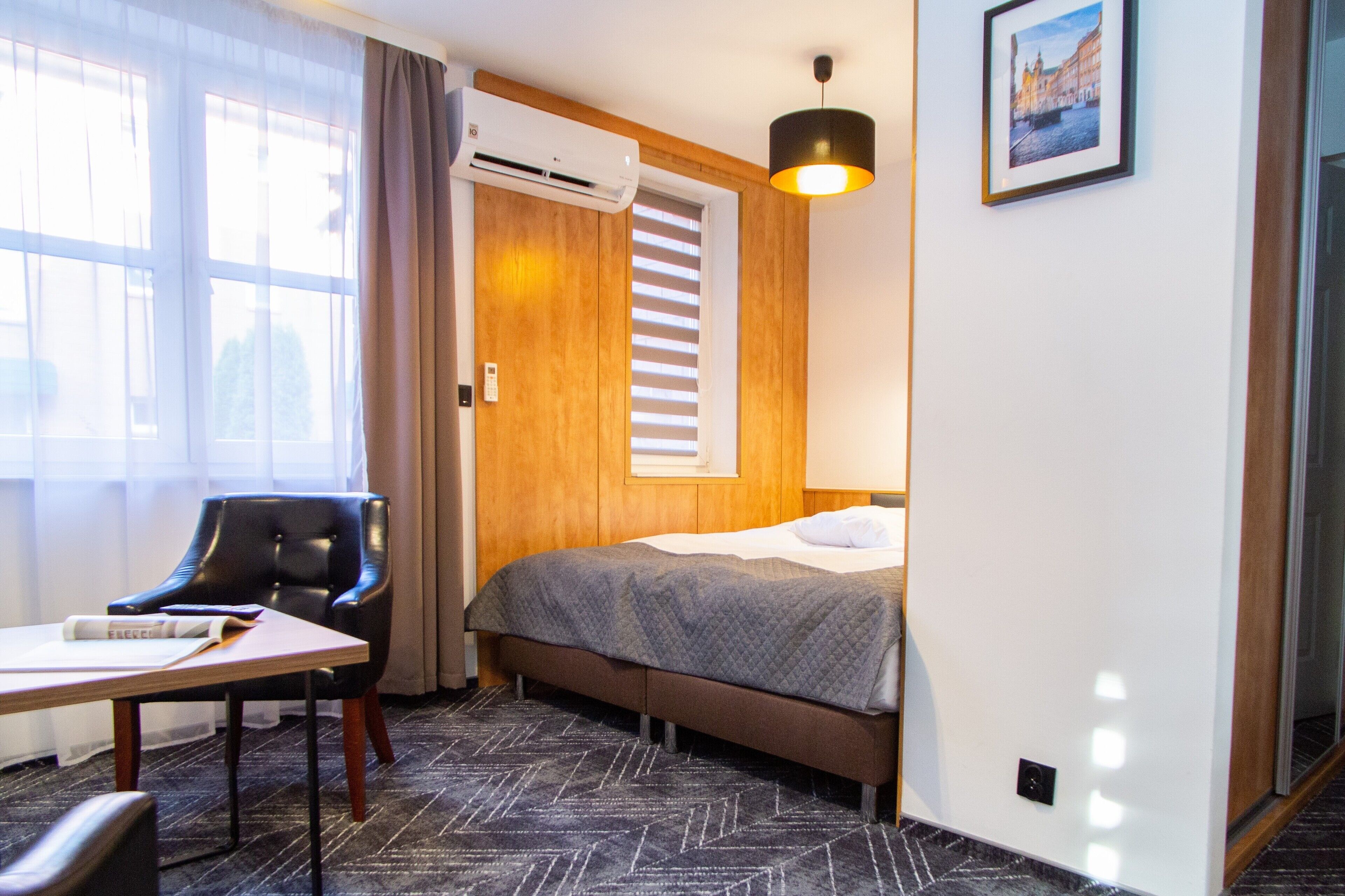 Habitación de hotel con cama, escritorio, silla, aire acondicionado, ventana con persianas y un cuadro en la pared.