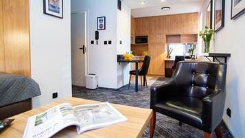 A modern hotel room with a wooden bed, a dining area with a table and chairs, a kitchen with wooden cabinets, and a black leather armchair.