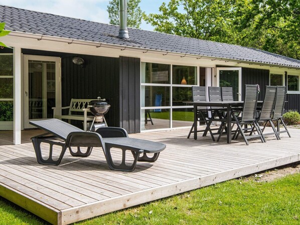 A covered outdoor patio with a table and chairs, a grill, and a deck extending onto a grassy area.
