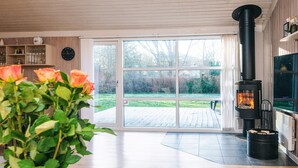 A modern living room with a large sliding glass door leading to a deck, a black wood-burning stove, and a green plant in the foreground. - Cozy Family Cottage - By Traum Ferienwohnungen (Hemmet)