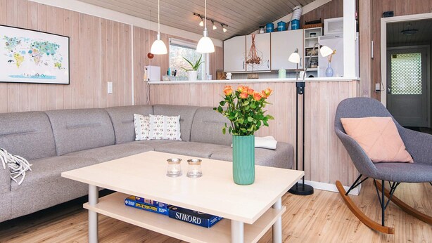 A modern living room with a sofa, a coffee table, and a rocking chair. There is a kitchen area with cabinets and a window. - Cozy Family Cottage - By Traum Ferienwohnungen (Hemmet)