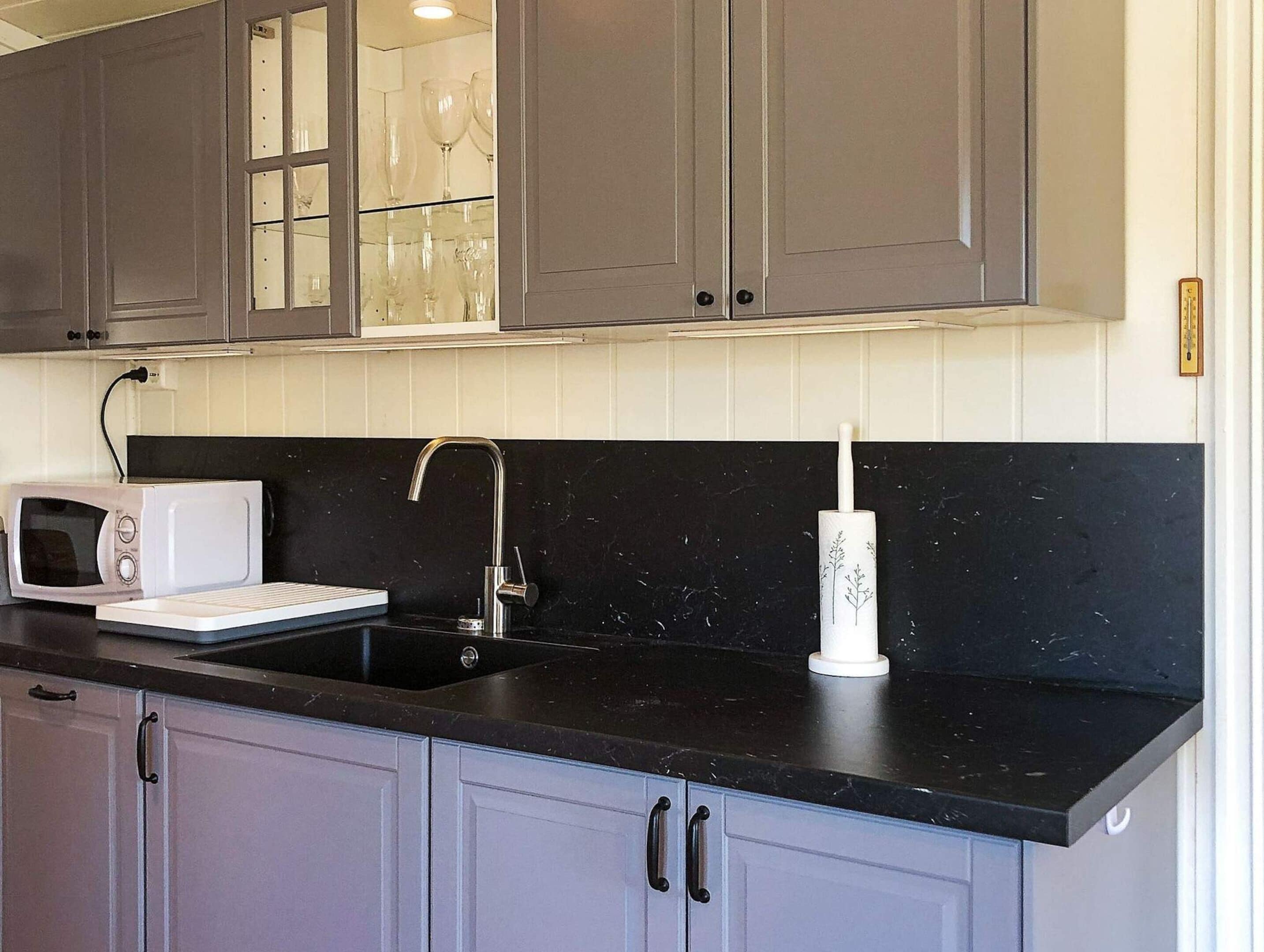 A kitchen with grey cabinets, a black countertop, a white microwave, a sink with a faucet, and a roll of paper towels.
