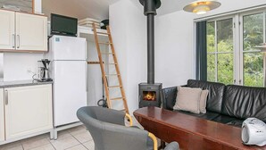 Et moderne køkken med hvide skabe, et køleskab, en kaffemaskine og en komfur. Der står en træstige lænet mod væggen, et spisebord med stole, og en sort lædersofa med en hvid højttaler.