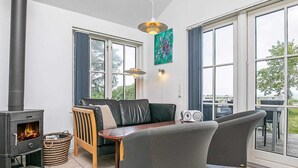 En hyggelig stue med en brændeovn, en sofa, et spisebord og store vinduer med udsigt til naturen.