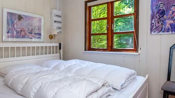 A bedroom with a bed, a window with a view of greenery, a wall-mounted electrical panel, and a framed painting.