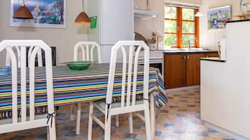 A kitchen with white cabinets, a white refrigerator, a stove, and a window with curtains. There is a table with chairs and a green bowl on it.