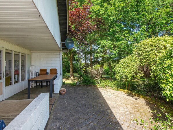 A covered patio with a table and chairs, a brick pathway, and a garden with trees and shrubs.