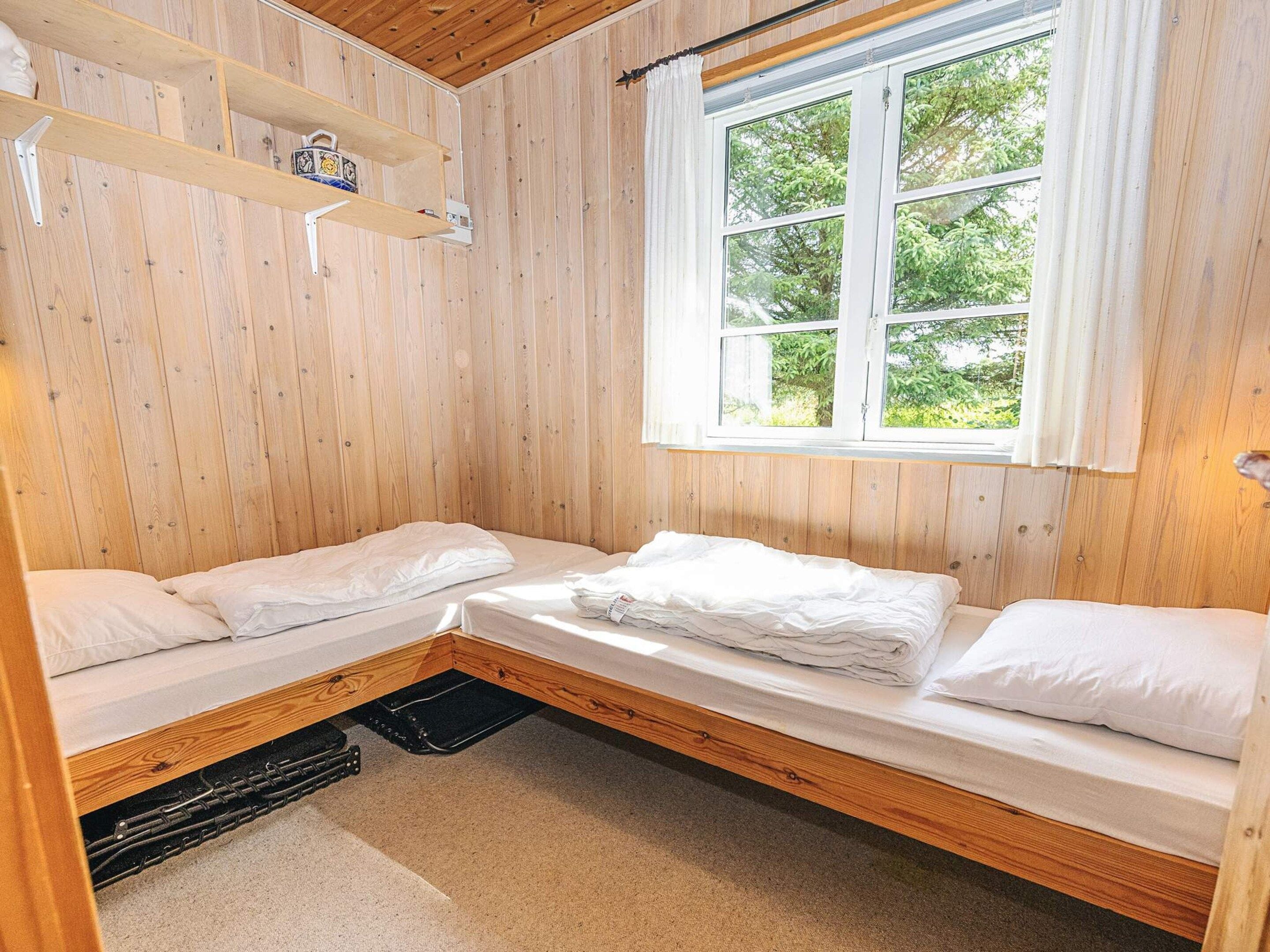 A small, cozy room with two single beds, a wooden ceiling, a window with a view of greenery, and a shelf with a decorative item.