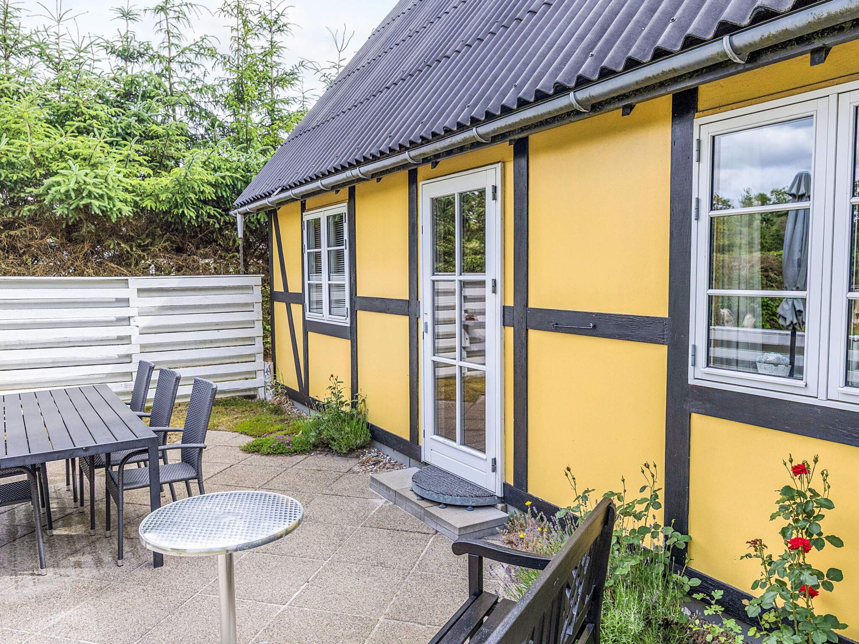 Un bâtiment jaune avec des bordures noires, une terrasse avec une table et des chaises, et un banc.