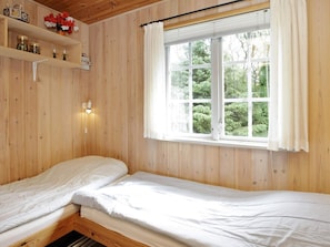 A room with wooden walls, a window with curtains, a bed, a shelf with jars, and a wall-mounted light.